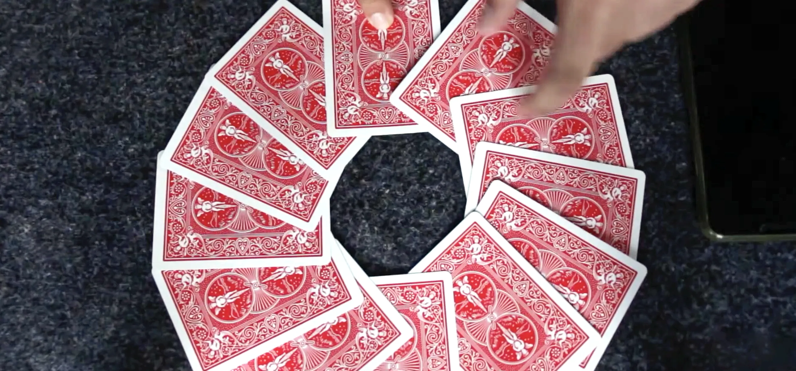 Hands execute 12 o'clock card trick, arranging red Bicycle playing cards in a circle on dark fabric, center hole highlighting clock illusion.