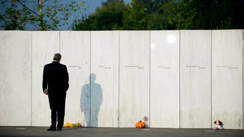 Man in a suit standing in front of a white memorial wall with flowers at the base.