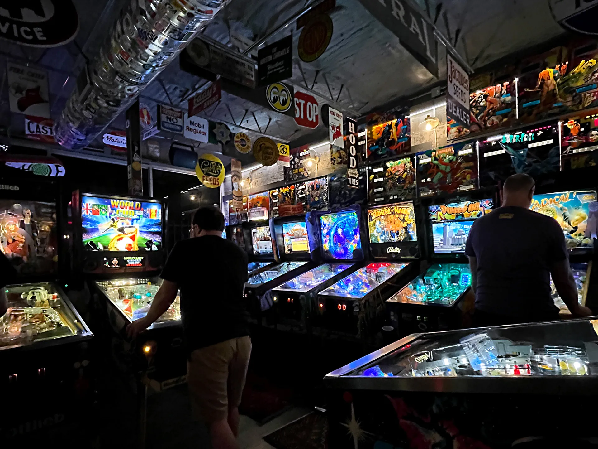 A dimly lit arcade featuring multiple pinball machines with vibrant screens and players interacting.