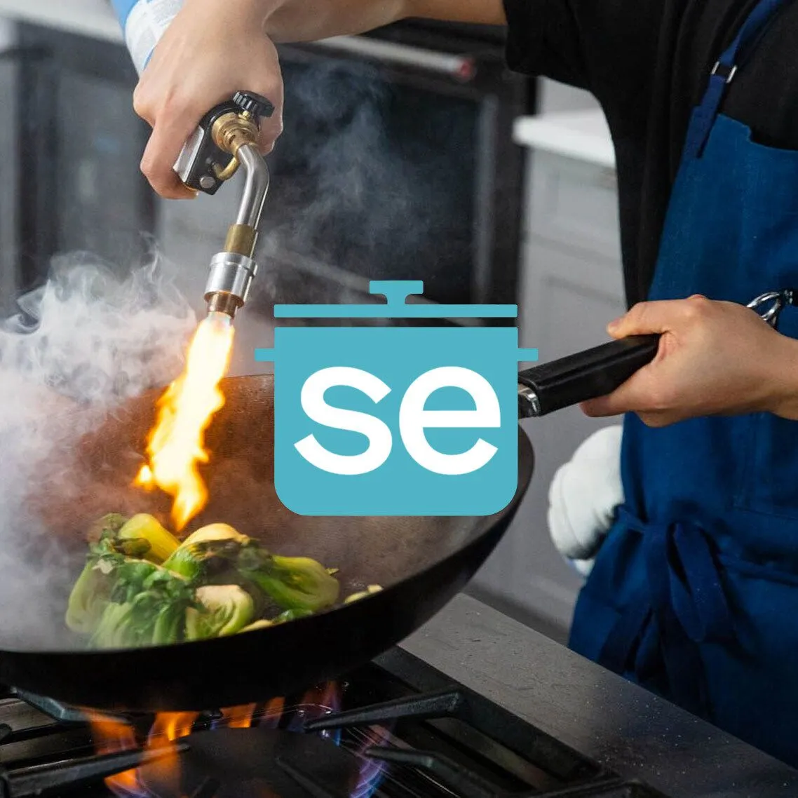 Chef using a torch to cook vegetables in a frying pan.