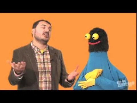 A man in a suit talking with a colorful puppet against an orange background.