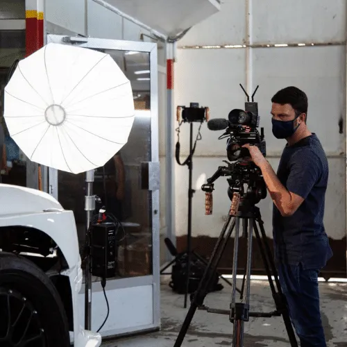 Video production setup featuring a camera operator in a mask, with lighting equipment and a vehicle in a studio environment.