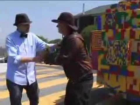 Two men wearing hats shake hands on a sunny street with a colorful mural in the background.