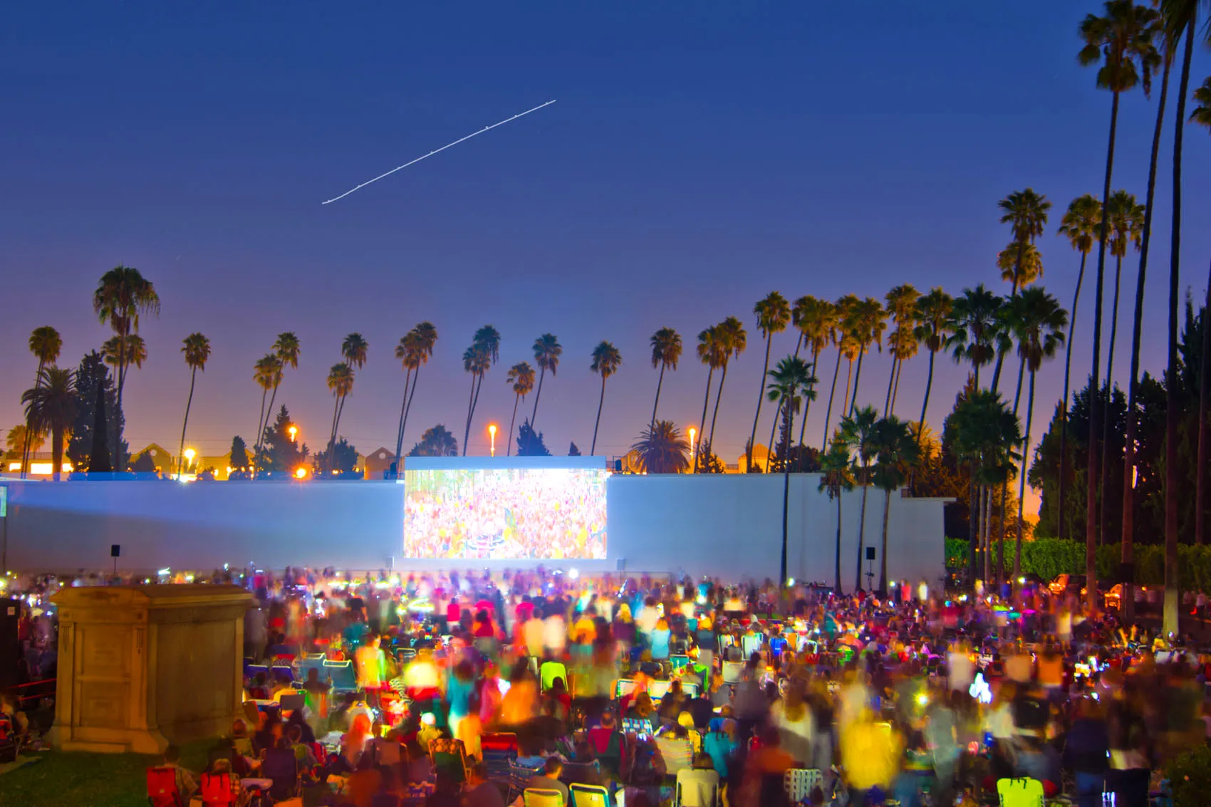 Outdoor movie screening with a large crowd under palm trees at twilight.