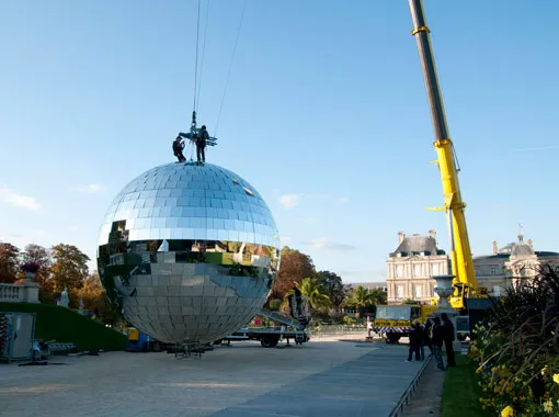 Workers installing a large reflective disco ball with a crane in an outdoor setting.