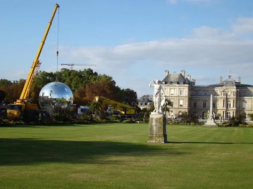 Cranes lifting a reflective sphere near a stately building and sculpture in a landscaped garden.