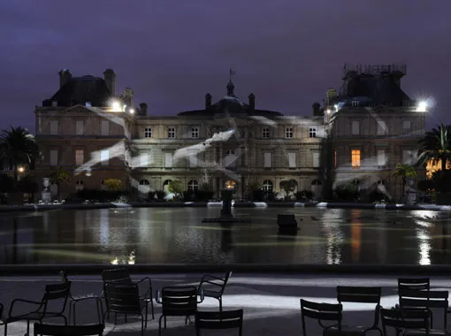 Historic building illuminated at night near a reflecting pool with chairs in the foreground.