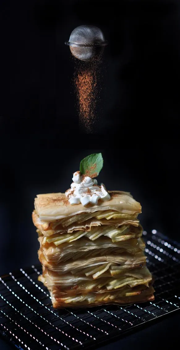 Dessert stack with layers of pastry, cream, and mint garnish against a dark background.