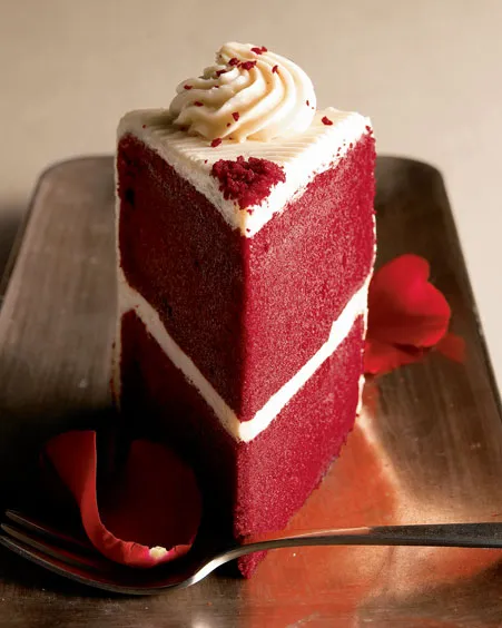 Red velvet cake slice with cream frosting and rose petals on a plate.