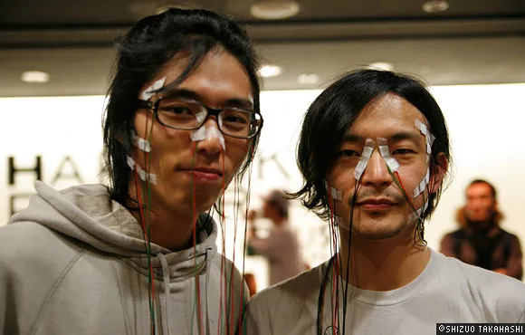 Two men with facial cream and colorful string accents on their faces, posing together.