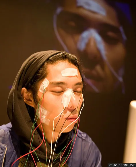 Person wearing a headscarf with electrodes on their face, engaged in a scientific experiment, with a projected face in the background.