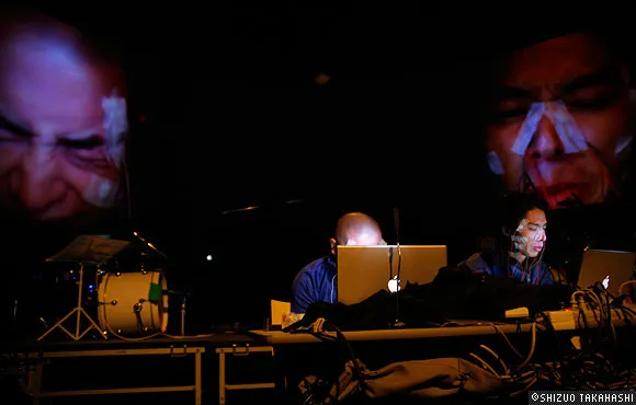 Musicians performing with laptops in a dark setting, featuring projected faces in the background.