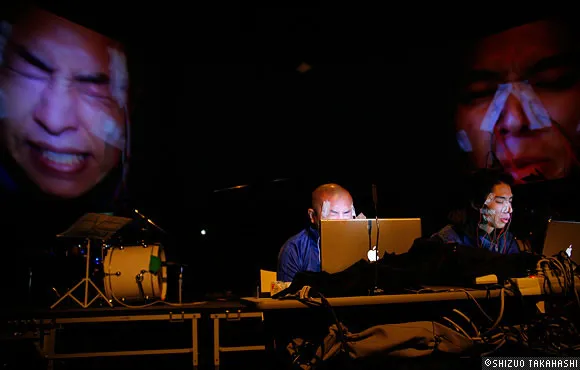 Performers using laptops on stage with projected expressive faces in the background.
