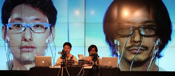 Two individuals performing on stage with large screens displaying faces behind them.