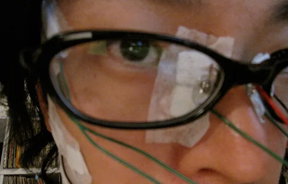 Person wearing glasses with medical tape and wires on their face.