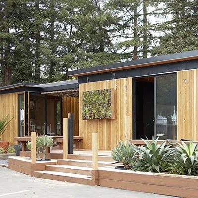 Modern wooden house with large windows and greenery in a forested area.