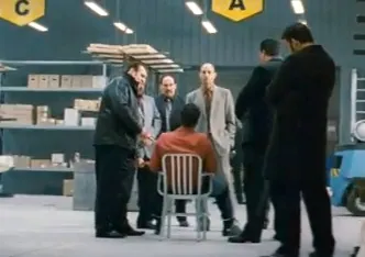 Group of men in suits discussing around a seated individual in a warehouse environment.