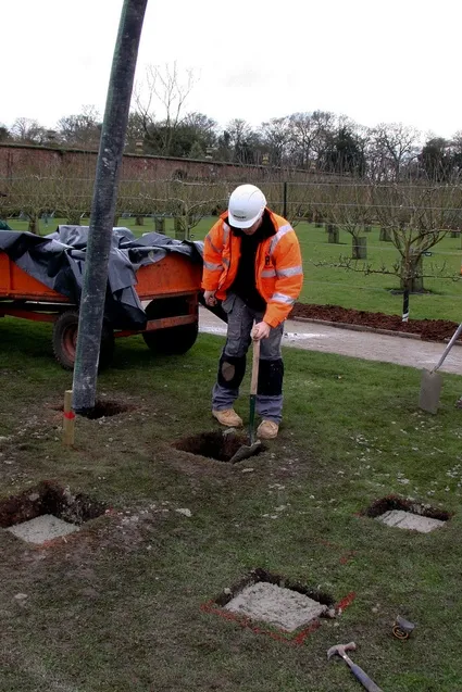 Worker digging a hole in a landscaped area with a pole and equipment nearby.