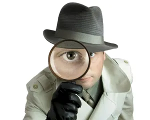 Man in a trench coat and fedora holding a magnifying glass up to his eye.