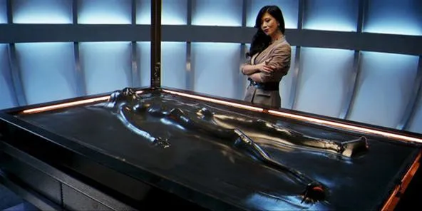 A woman observing a humanoid figure on a sleek, futuristic table in a laboratory setting.