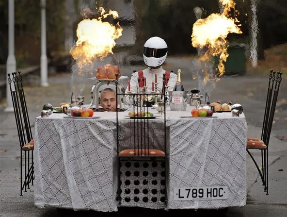A humorous scene featuring a person in a racing suit and helmet sitting at a dining table on a modified vehicle, with flames shooting upward.