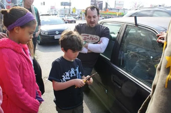 Child holding a tool near a car with adults observing.