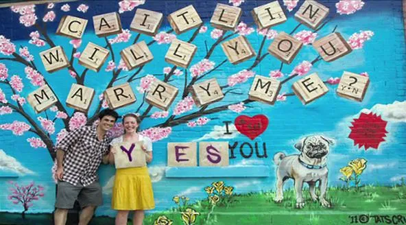 Couple posing in front of a colorful mural asking "Caitlin, will you marry me?" with playful decorations.