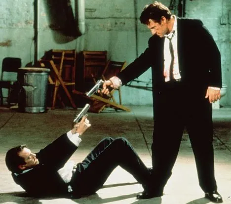 Confrontation between two men in suits, one holding a gun while the other is on the ground.