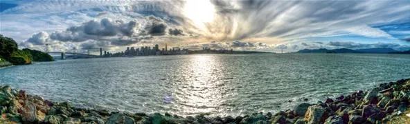 Panoramic view of a tranquil waterfront with city skyline in the background under a dramatic sky.