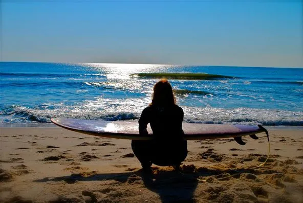 Person sitting on the beach with a surfboard, gazing at the ocean.