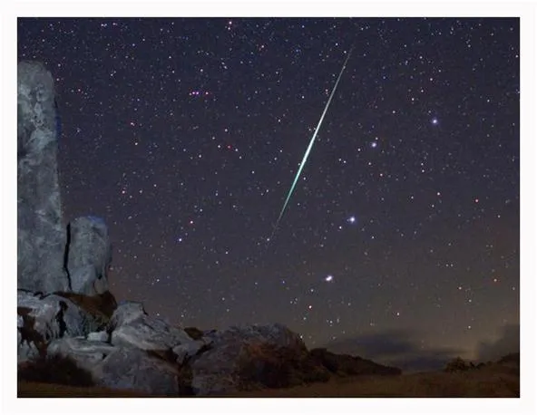 Night sky with stars and a meteor streaking over rocky landscape.