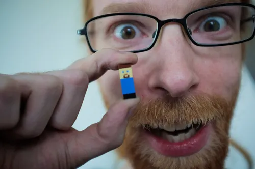 A man with glasses holds a small Lego figure, smiling enthusiastically.