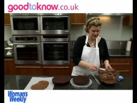 Woman spreading icing on a chocolate cake in a modern kitchen.