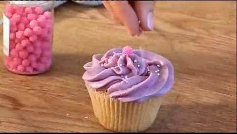 Cupcake with purple frosting being decorated with pink sprinkles.