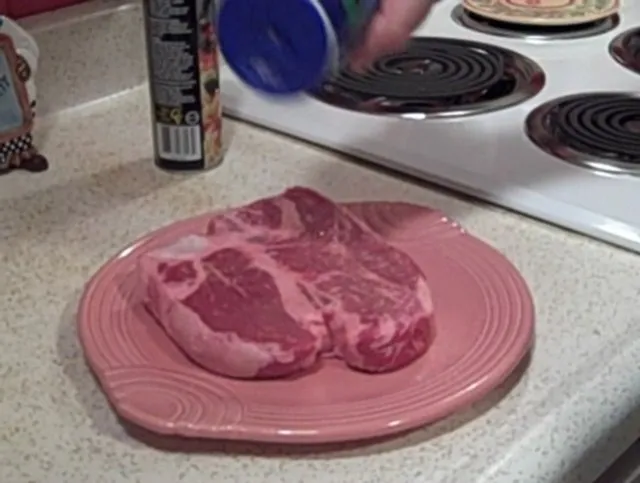 Raw steak being seasoned on a pink plate in a kitchen setting.