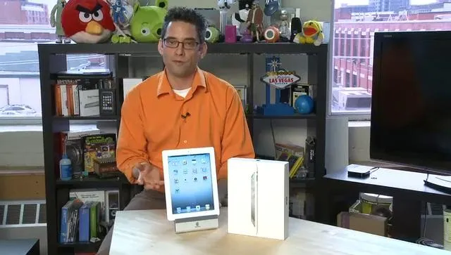 A person in an orange shirt presenting an iPad and its packaging in an office setting.