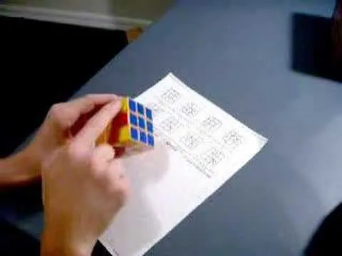 A person solving a Rubik's Cube with a reference sheet on the table.
