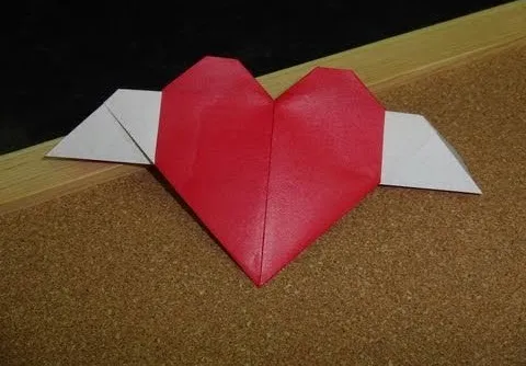 Red origami heart with white wings on a cork surface.