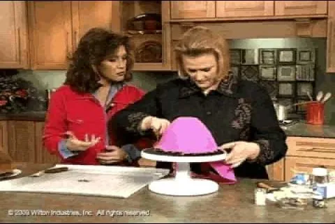 Two women decorating a cake with a pink cover in a kitchen setting.