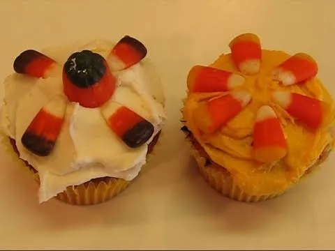 Two decorative cupcakes with candy toppings, one featuring a white frosting with candy corn and a dark candy center, and the other with orange frosting and candy corn arranged in a floral pattern.