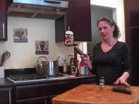 Woman in a kitchen holding a cucumber and standing by a cooking pot.