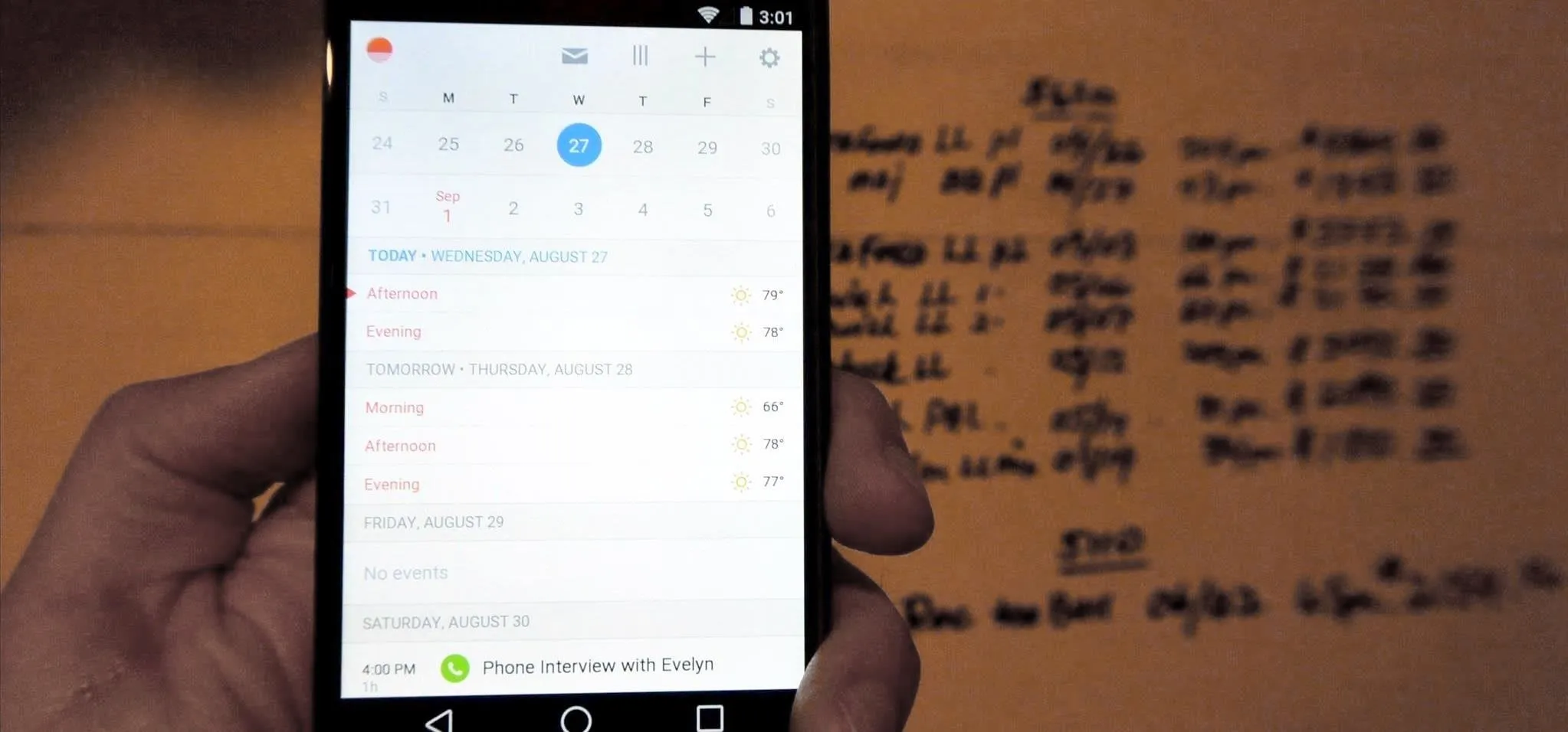 A hand holding a smartphone displaying a calendar app next to a wall with handwritten notes.