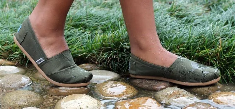 Woman wearing green flats stepping on stones in a grassy area.