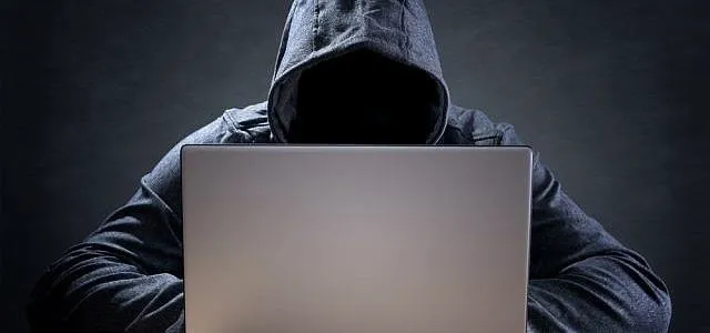 Hooded figure using a laptop in a dimly lit environment.