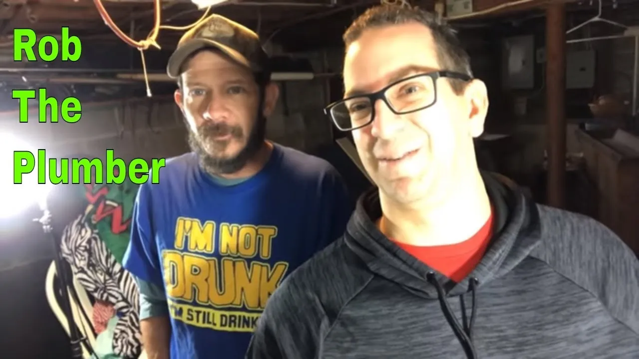 Two men smiling in an indoor setting, one wearing a blue shirt and the other in a gray hoodie, with text identifying one as "Rob The Plumber."