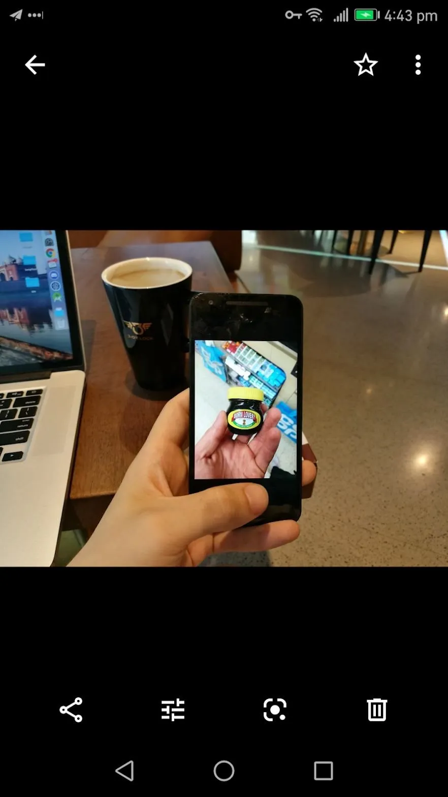 A hand holding a smartphone displaying a fitness tracking app, with a laptop and coffee cup in the background.