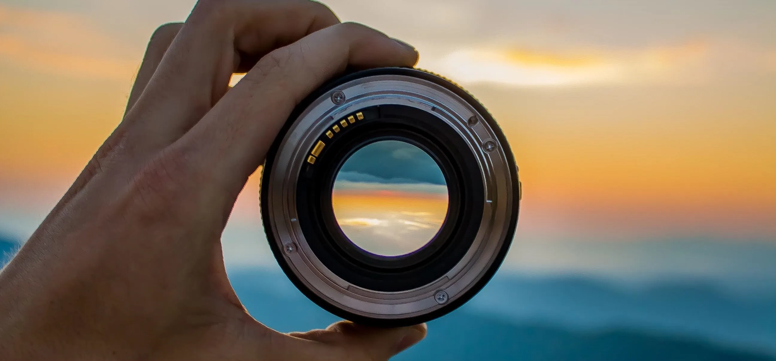 A hand holding a camera lens with a scenic sunset in the background.