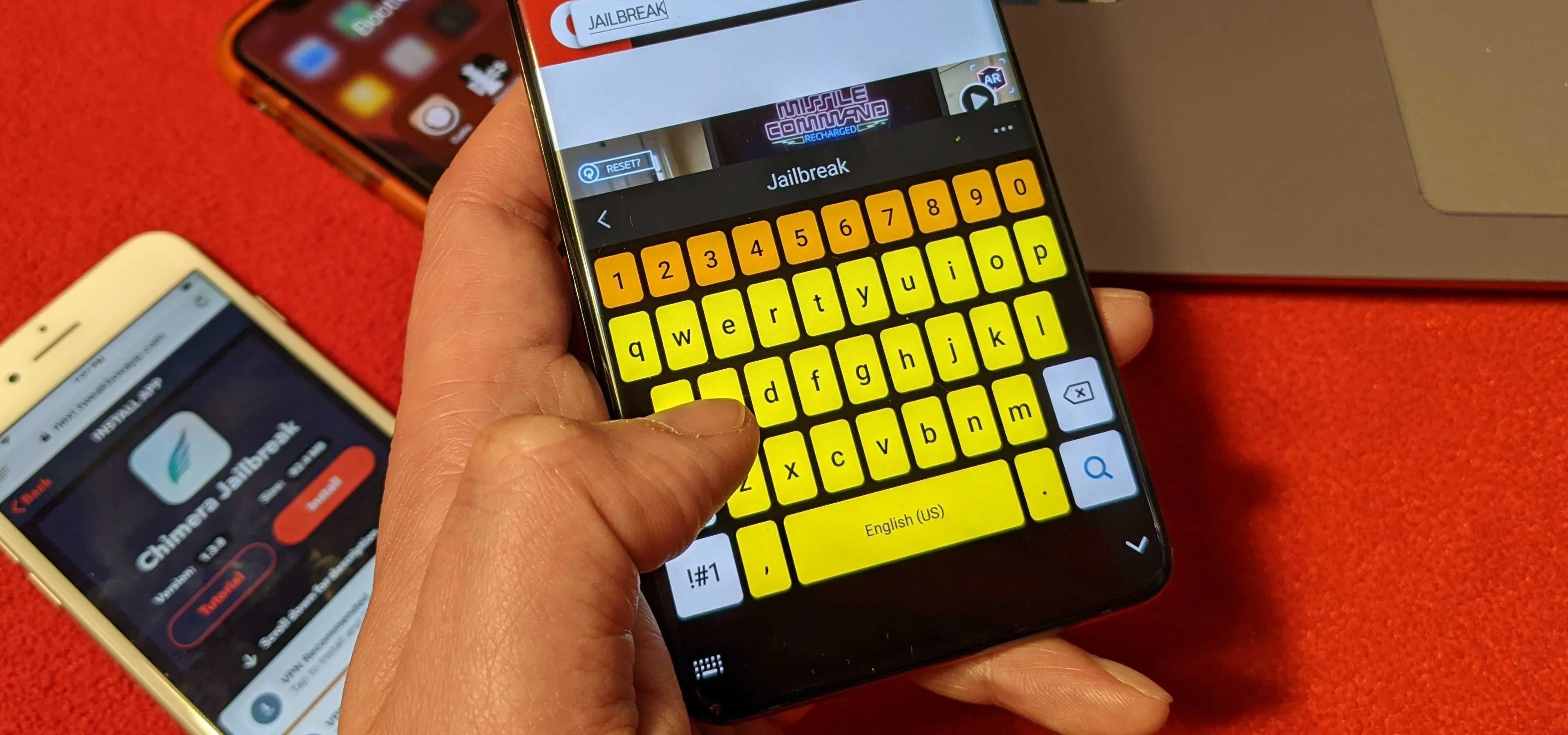 Person using a smartphone with a colorful keyboard on a red surface, with another phone and a laptop in the background.