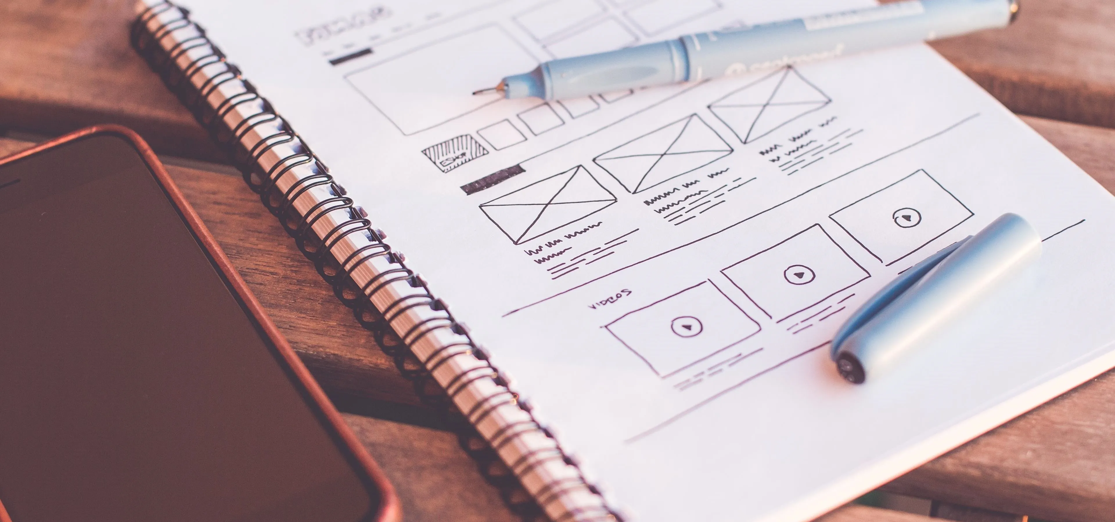 Notebook with wireframe sketches, a pen, and a smartphone on a wooden surface.