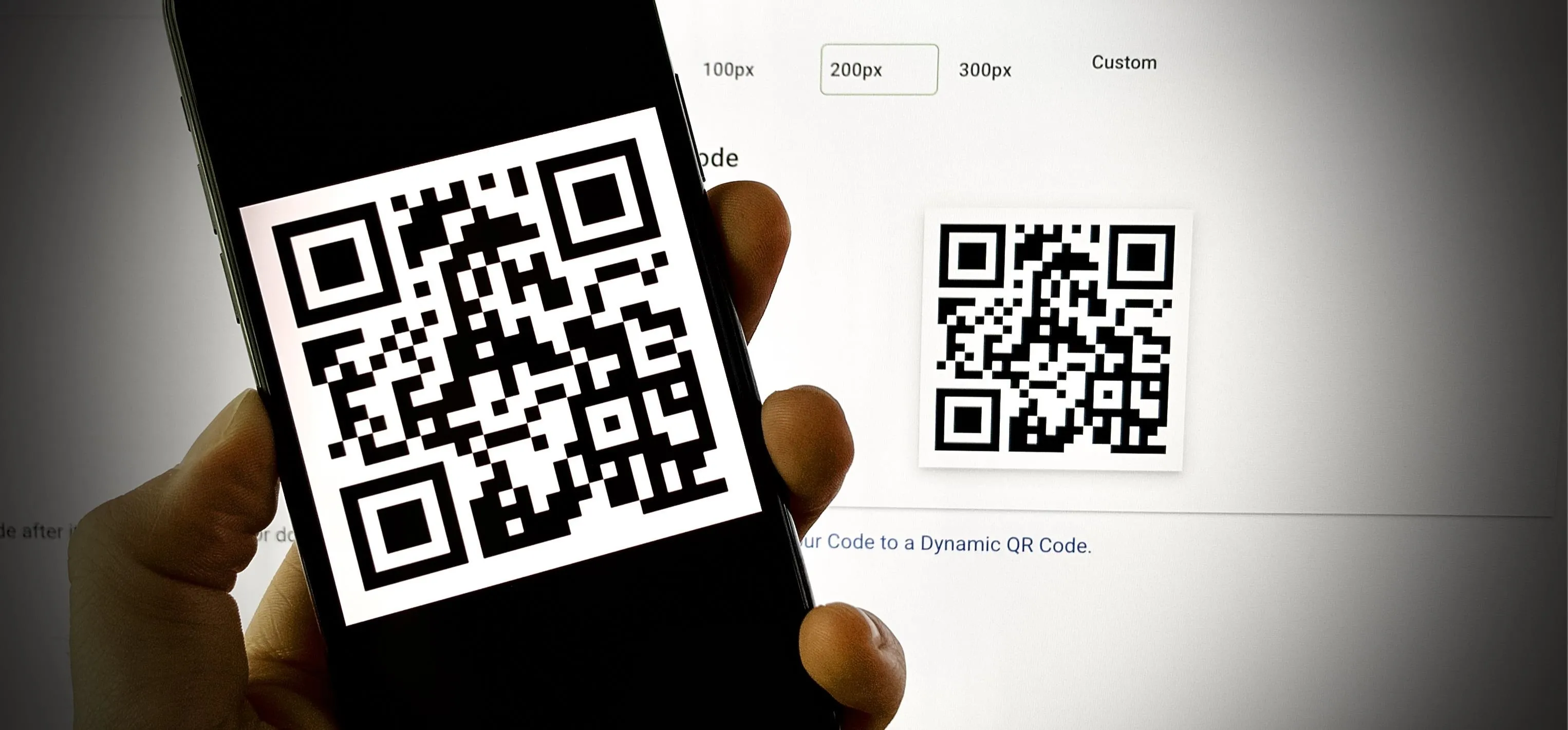 A hand holding a smartphone displaying a QR code, with another QR code shown in the background.